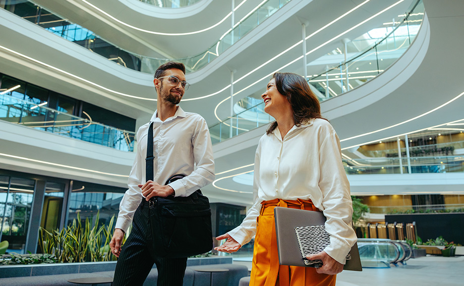 In the lobby of a modern building, a man with glasses gazes at a woman, who is smiling and speaking to him.