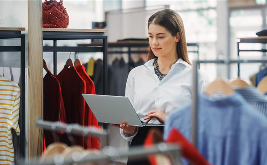 A woman stands among clothes, operating a laptop in a clothing store