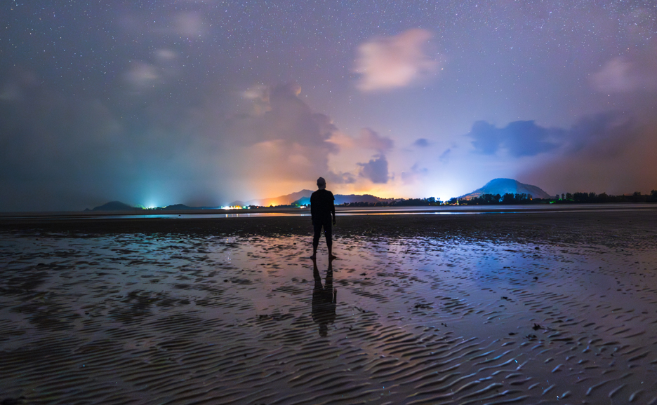 Silhouette of a person standing on a wet beach at night, with city lights in the distance.