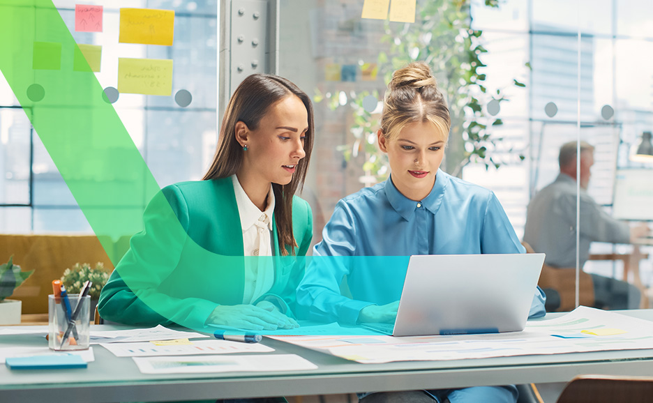 Two women collaborating in a bright office, with one woman in a green jacket pointing at the laptop screen of the woman next to her.